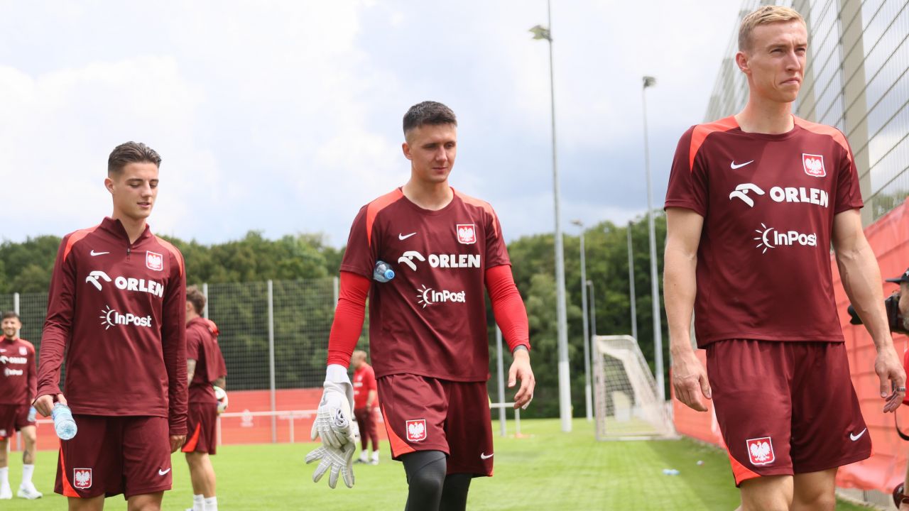 Trzech zawodników reprezentacji Polski podczas treningu na boisku. W centrum kadru Mateusz Kochalski — bramkarz w bordowym stroju treningowym z rękawicami w dłoni. Zawodnik jest obserwowany przez Borussię Dortmund po świetnych występach w barwach Karabachu Agdam, z którym awansował do Ligi Mistrzów. Trening odbywa się w słoneczny dzień na profesjonalnym obiekcie sportowym, w tle pozostali zawodnicy oraz bramki (fot. Getty).