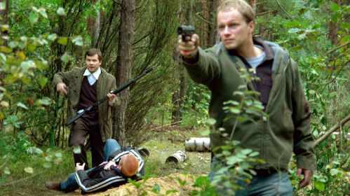 Serial „Glina” – surowy realizm, mrok i niejednoznaczni bohaterowie – oglądaj wTVP VOD. Dwóch mężczyzn z bronią w lesie, jeden celuje, drugi stoi przy leżącym człowieku.